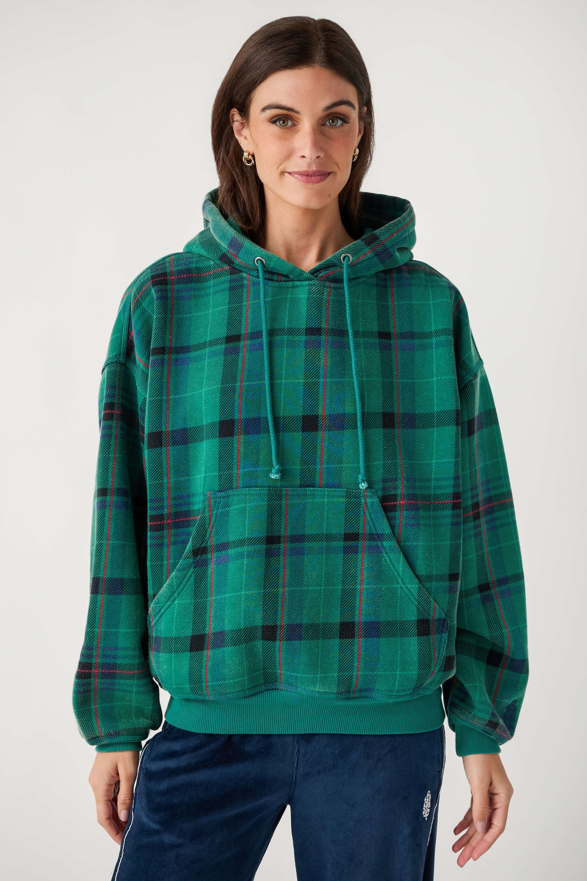 A woman wears a green plaid hooded sweatshirt with red and black stripes, a front pouch pocket, and navy velour pants.