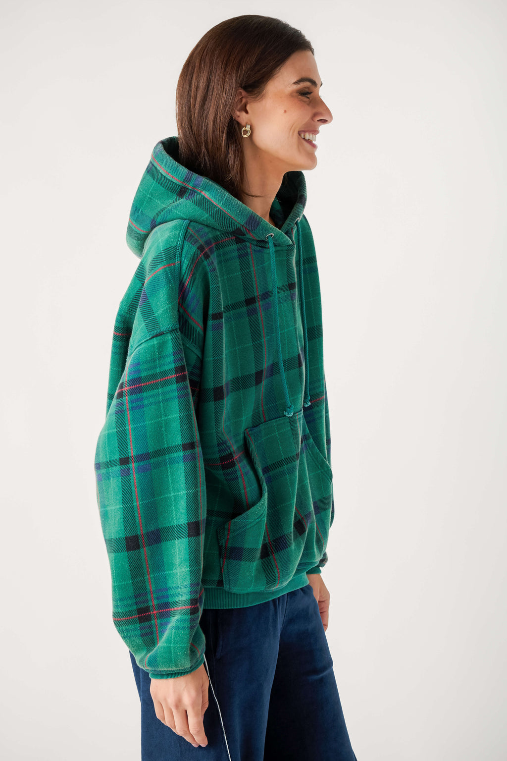 A smiling woman in profile wears an oversized green plaid hooded sweatshirt with a front pocket and navy velour pants.