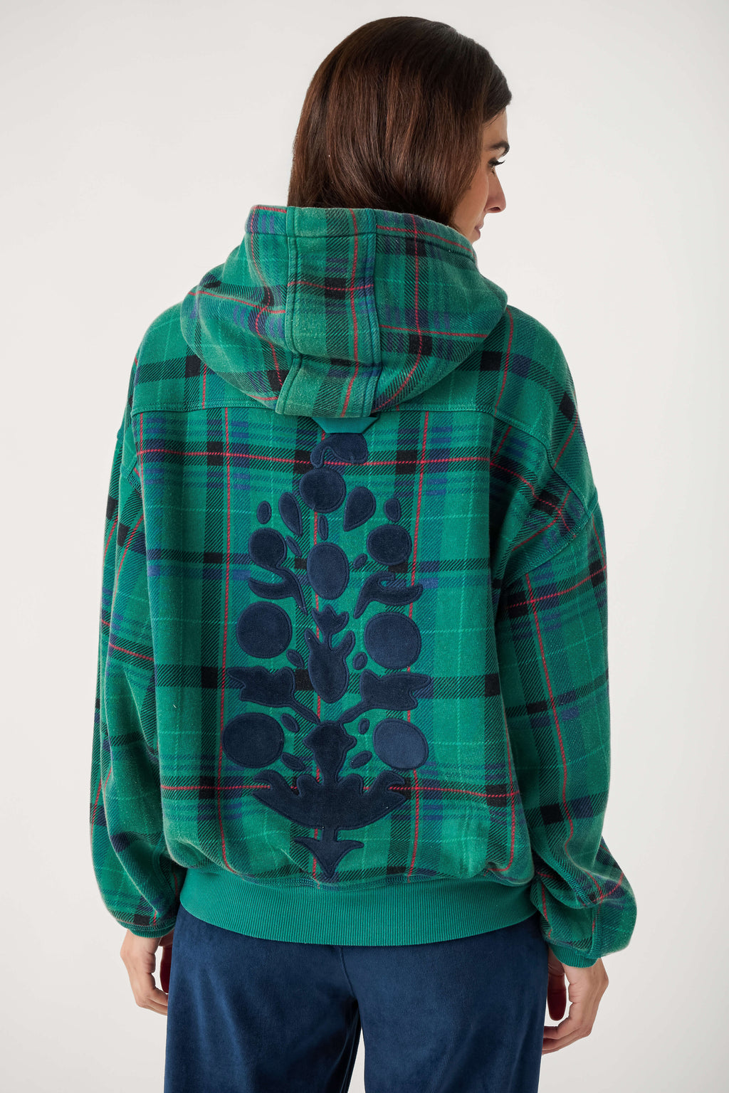 Back view of a woman wearing a green plaid hooded sweatshirt with a navy floral appliqué down the back and navy velour pants.