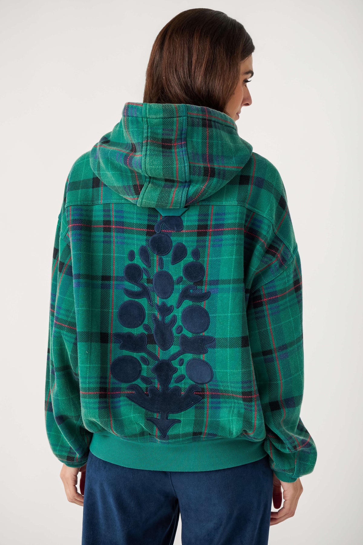 Back view of a woman wearing a green plaid hooded sweatshirt with a navy floral appliqué down the back and navy velour pants.