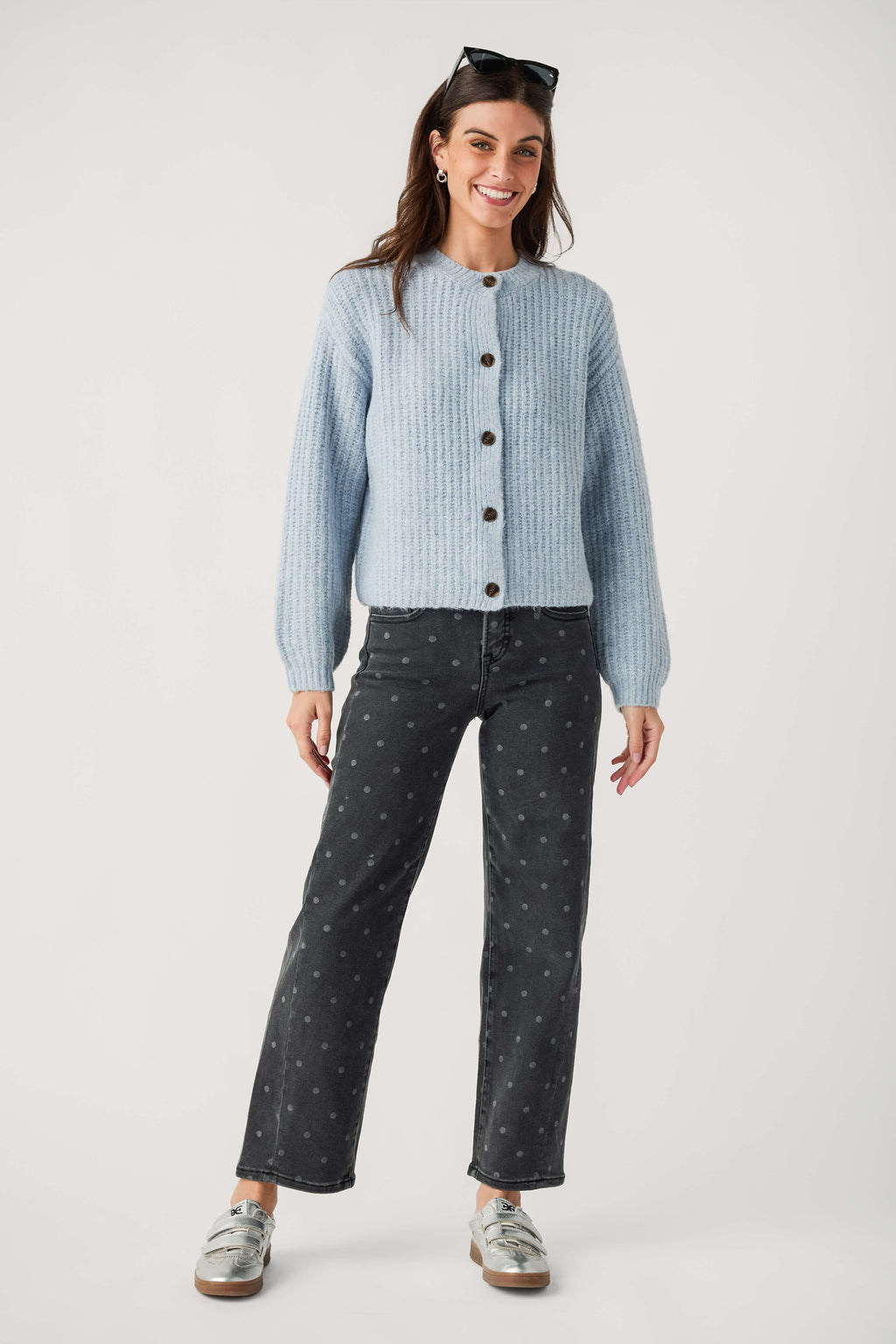 A smiling woman wears faded black polka dot ankle jeans with a light blue knit cardigan and silver sneakers.