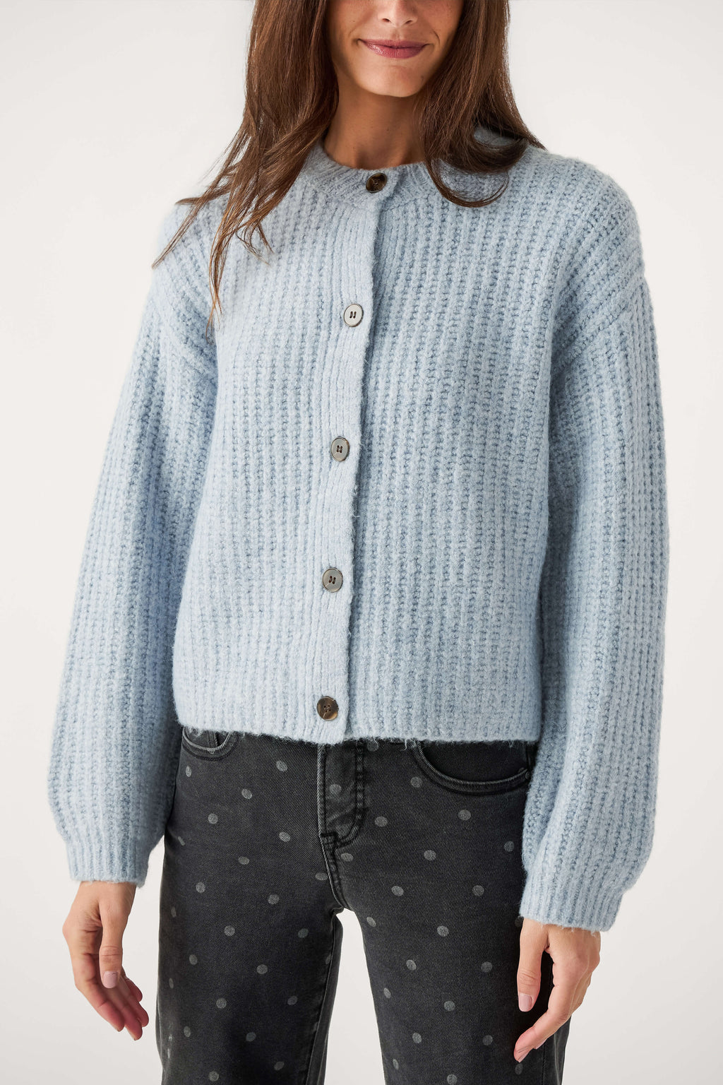 A woman wears a light blue ribbed knit cardigan with dark buttons and gray polka dot black jeans.