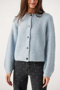 A woman wears a light blue ribbed knit cardigan with dark buttons and gray polka dot black jeans.