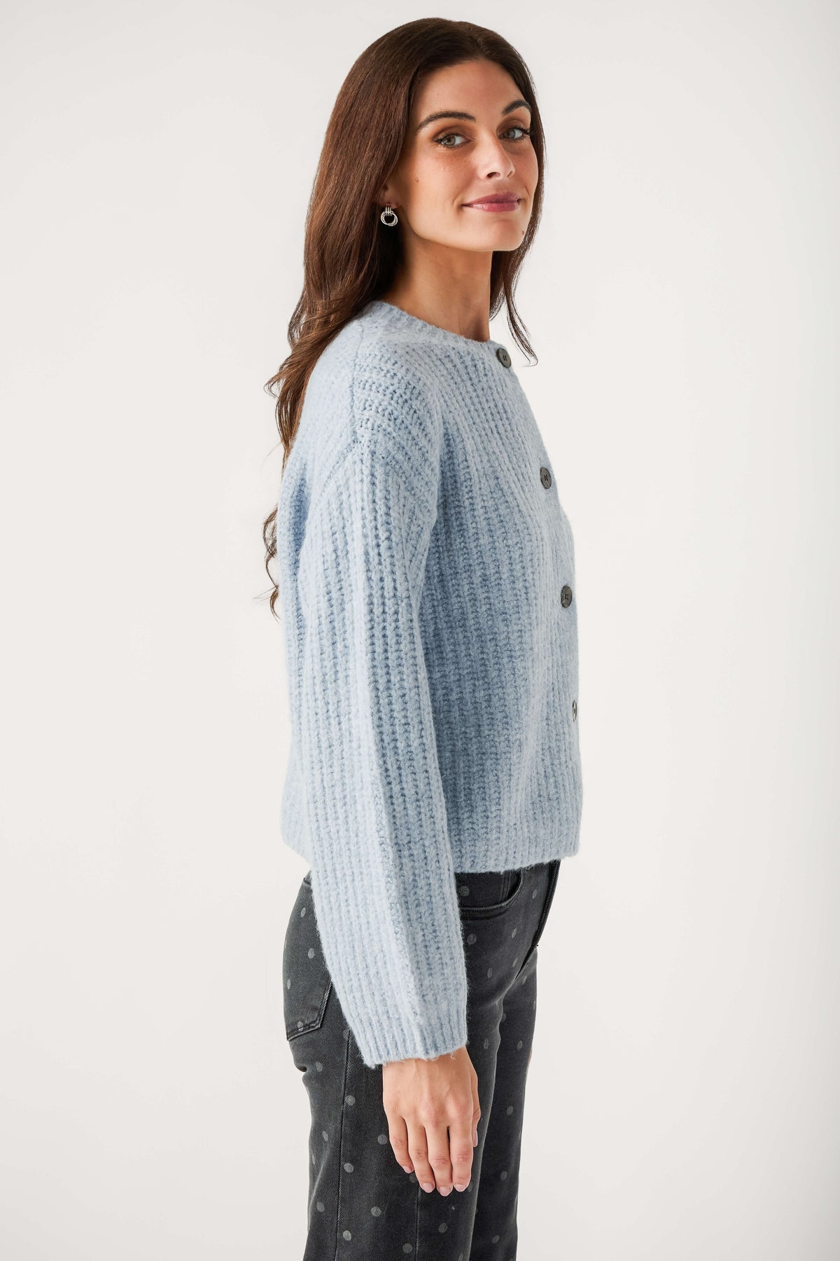 A woman with long brown hair models a light blue chunky knit cardigan with dark buttons and polka dot gray jeans.
