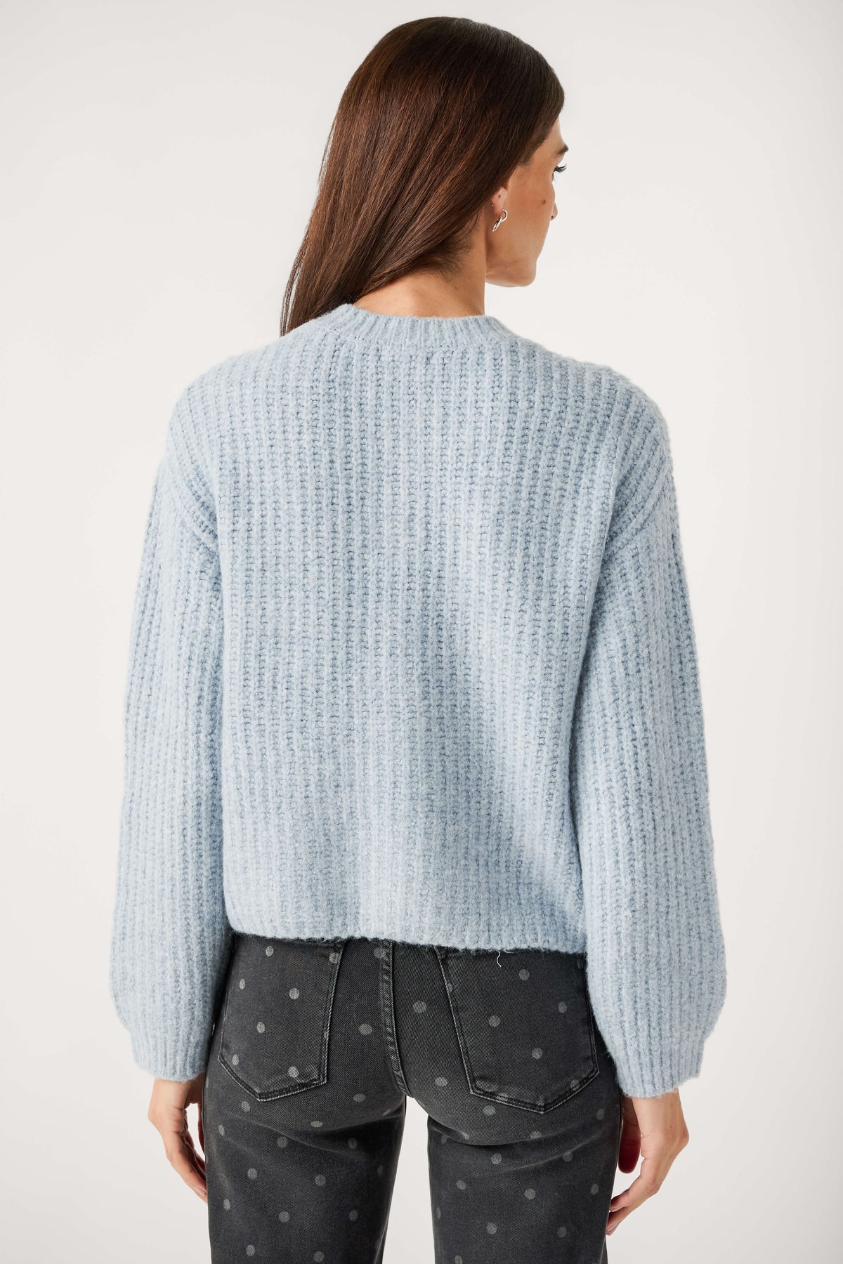 Back view of a woman wearing a light blue ribbed knit cardigan with full sleeves and dark gray polka dot jeans.