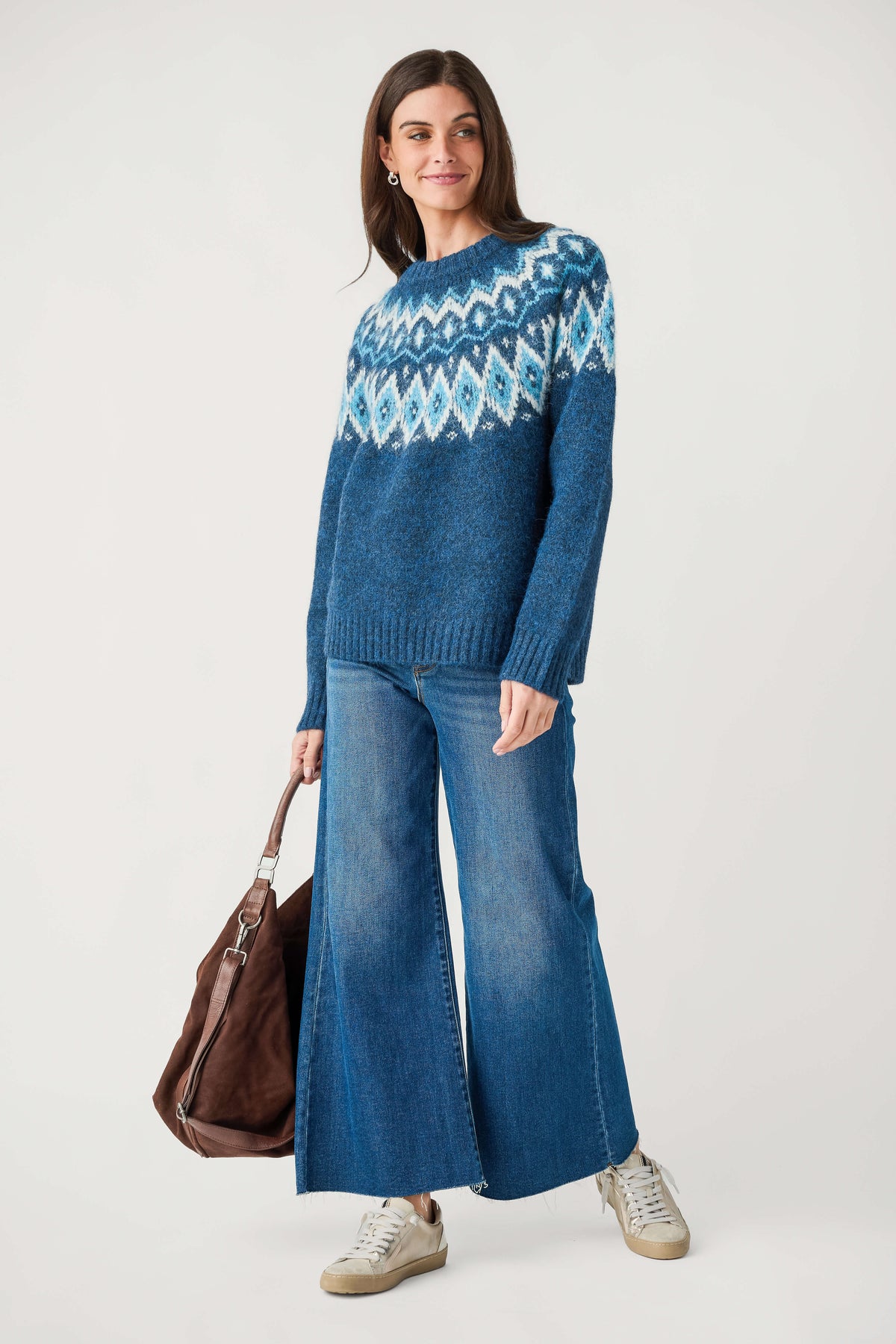 A woman wears a blue crewneck sweater with a white and light blue Fair Isle yoke, wide-leg jeans, and holds a brown suede bag.