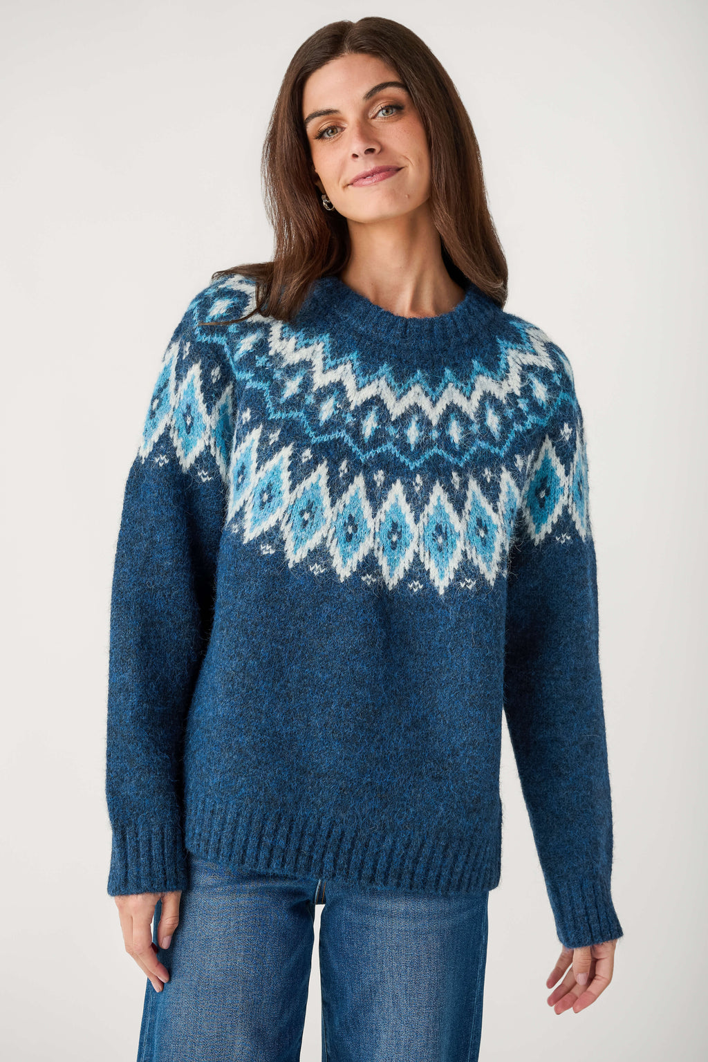 A woman wearing a blue Fair Isle knit sweater with geometric patterns in white and light blue, paired with blue jeans, against a plain light gray background.