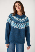 A woman wearing a blue Fair Isle knit sweater with geometric patterns in white and light blue, paired with blue jeans, against a plain light gray background.
