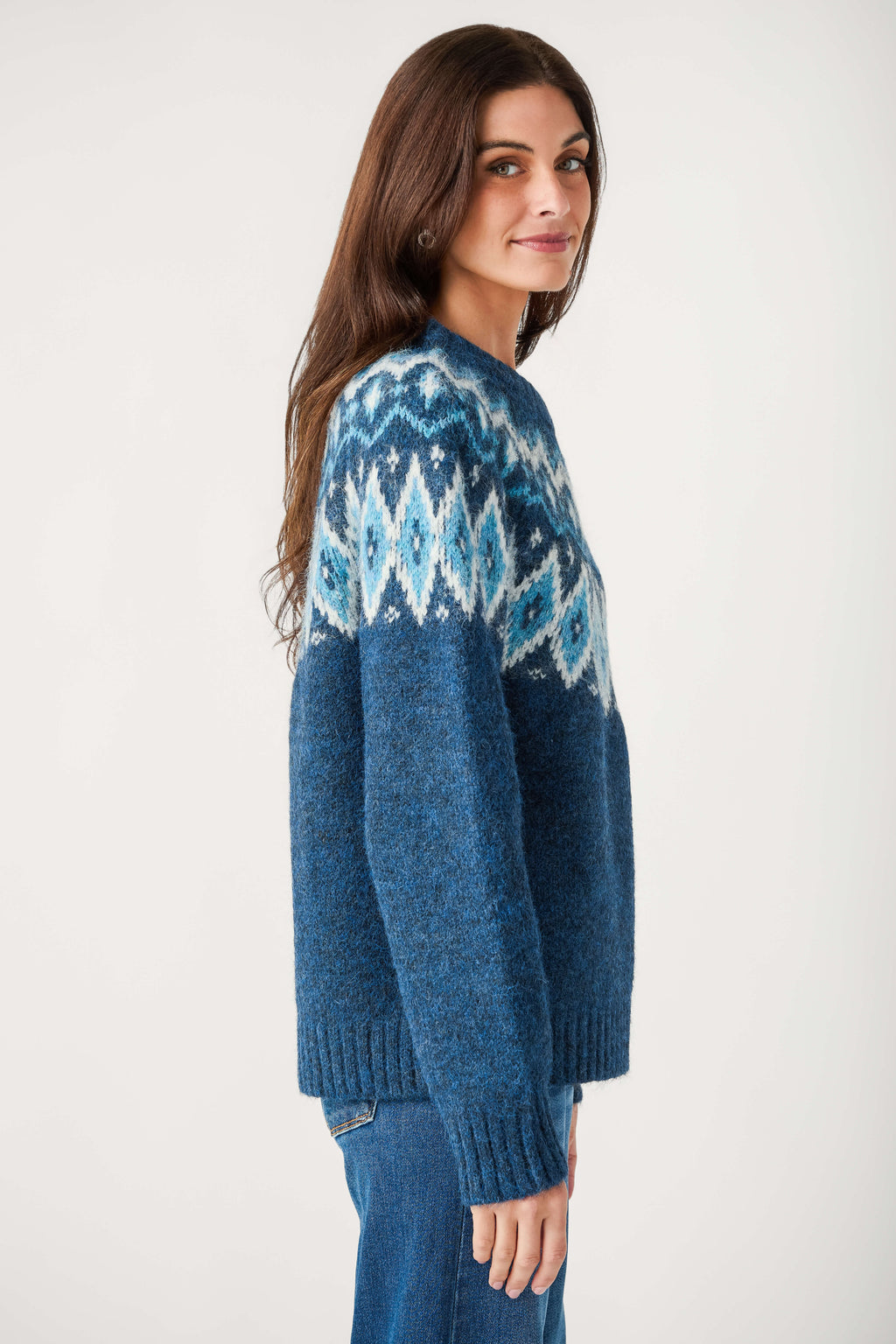 A woman with long brown hair wears a blue crewneck sweater with a white and light blue Fair Isle yoke and blue jeans.