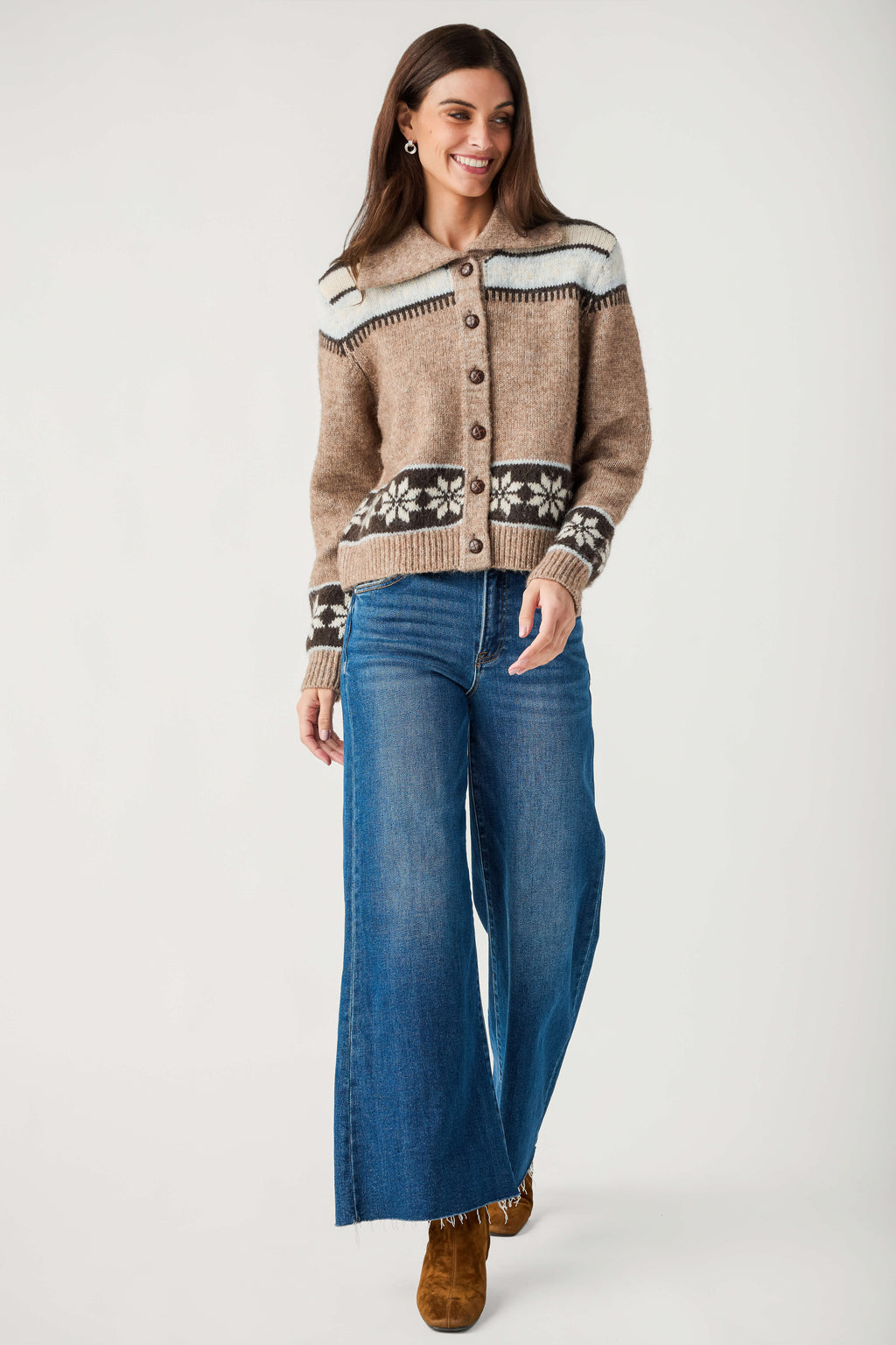 A smiling woman wears a tan button-front cardigan with snowflake pattern trim, wide-leg blue jeans, and brown suede ankle boots.