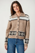 A woman wearing a cropped knit cardigan sweater with a brown base, cream and dark brown geometric patterns on the yoke and hem, and dark brown snowflake motifs on the cuffs, paired with blue jeans against a light gray studio background.