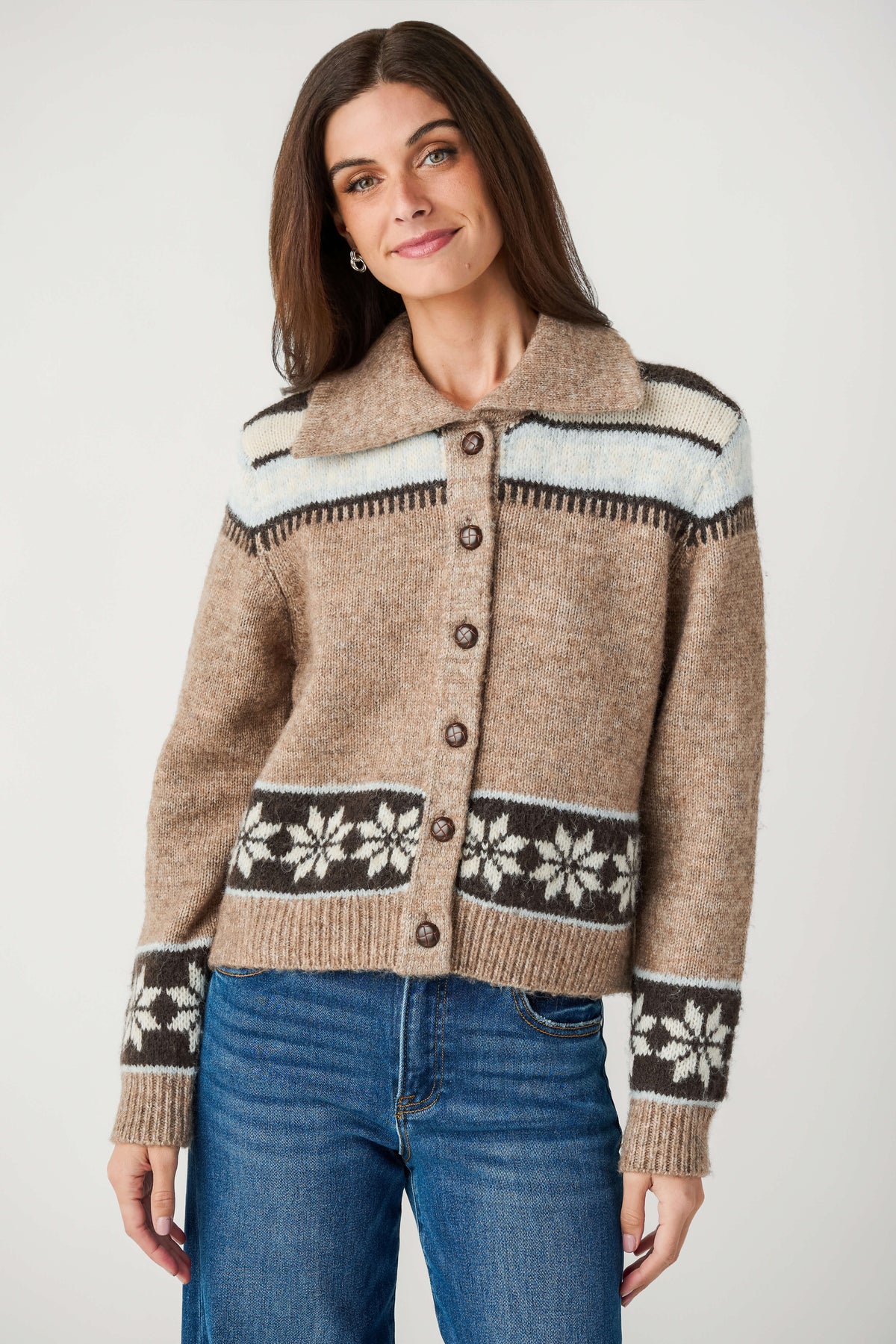 A woman wearing a cropped knit cardigan sweater with a brown base, cream and dark brown geometric patterns on the yoke and hem, and dark brown snowflake motifs on the cuffs, paired with blue jeans against a light gray studio background.