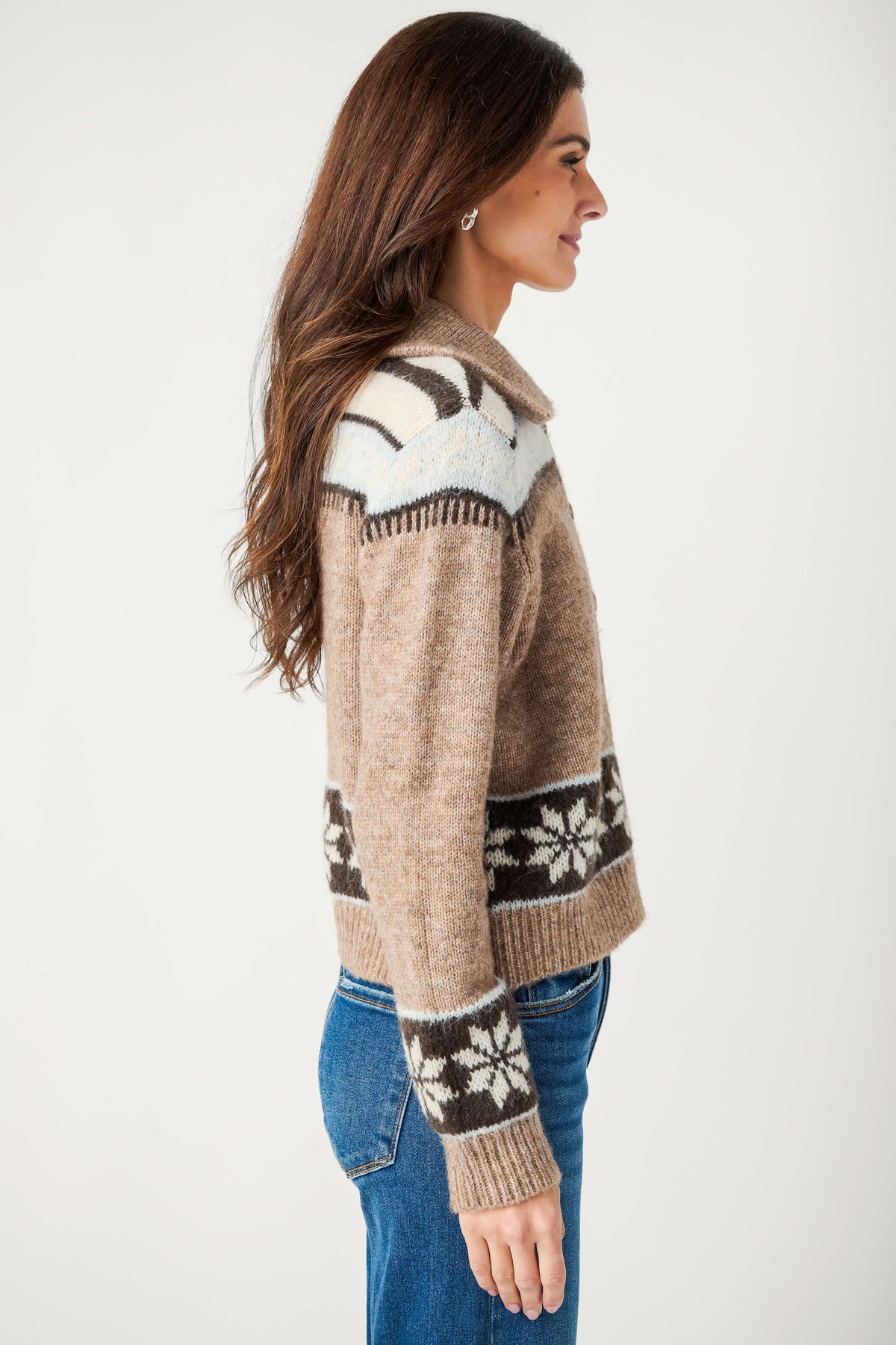 A woman in profile wears a tan knit collared cardigan with brown and cream snowflake bands and light blue yoke, paired with blue jeans.