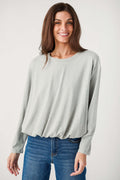 A smiling woman wears a heather gray long-sleeve top with a gathered hem, styled with blue denim jeans.