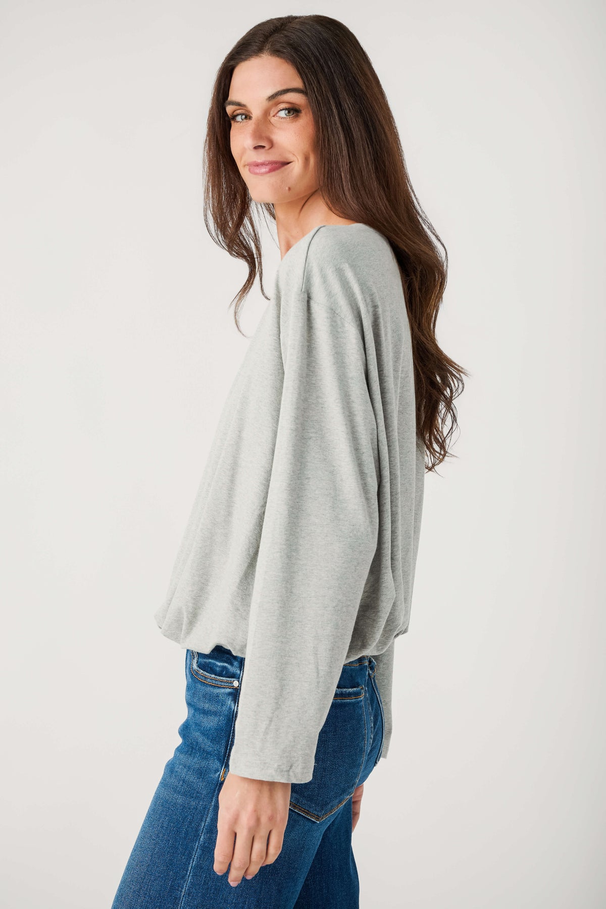 A woman with long brown hair wears a heather gray long-sleeve top with a gathered hem and blue jeans, turned in profile.