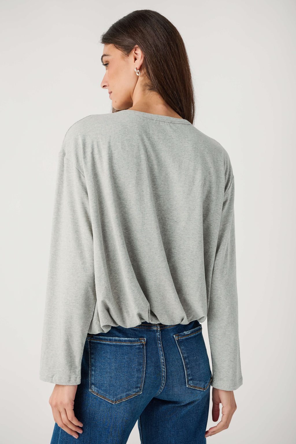 A woman seen from behind wears a heather gray long-sleeve top with a gathered hem and blue jeans.
