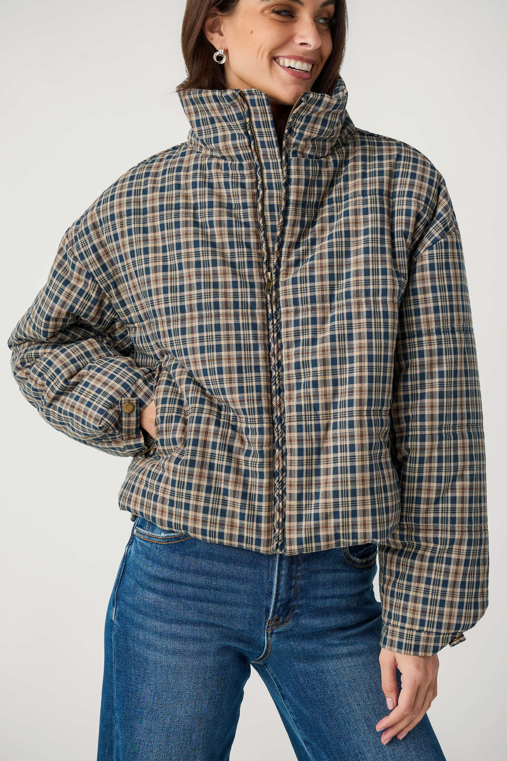 A smiling woman wears a navy, tan and brown plaid puffer jacket with a high collar over blue jeans.