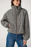 A smiling woman wears a navy, tan and brown plaid puffer jacket with a high collar over blue jeans.