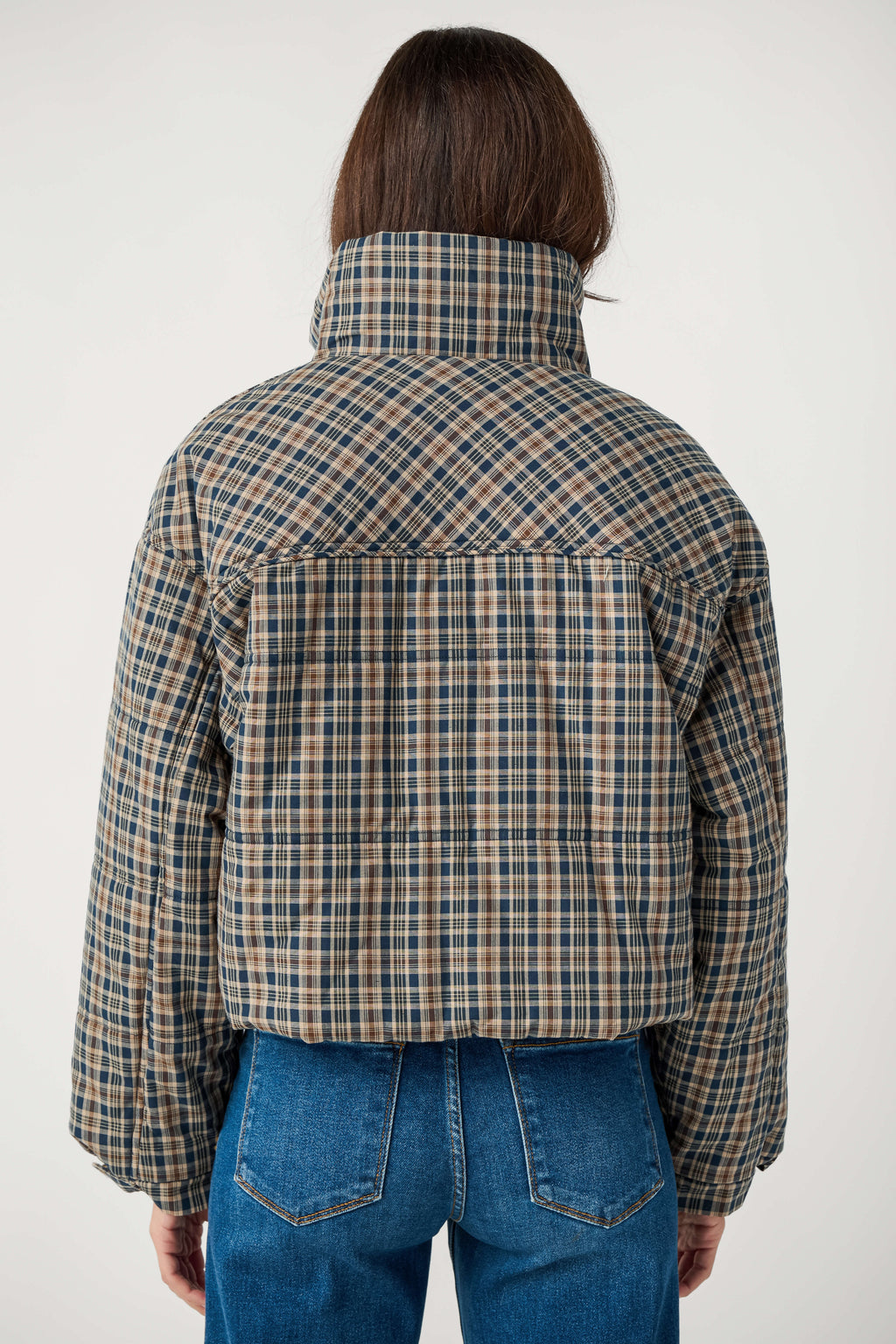 Back view of a person wearing a cropped navy, tan and brown plaid puffer jacket with a high collar and blue jeans.