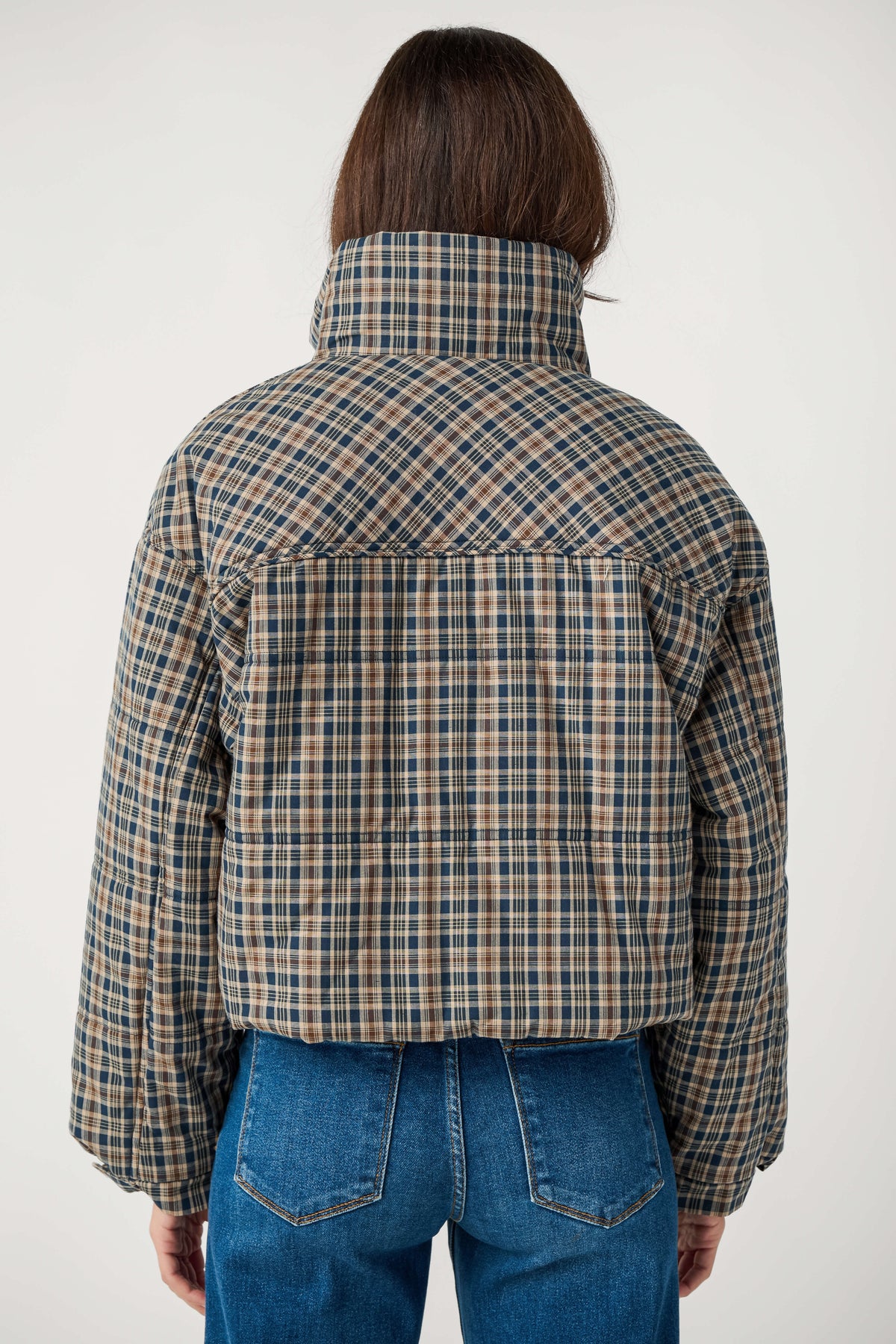 Back view of a person wearing a cropped navy, tan and brown plaid puffer jacket with a high collar and blue jeans.