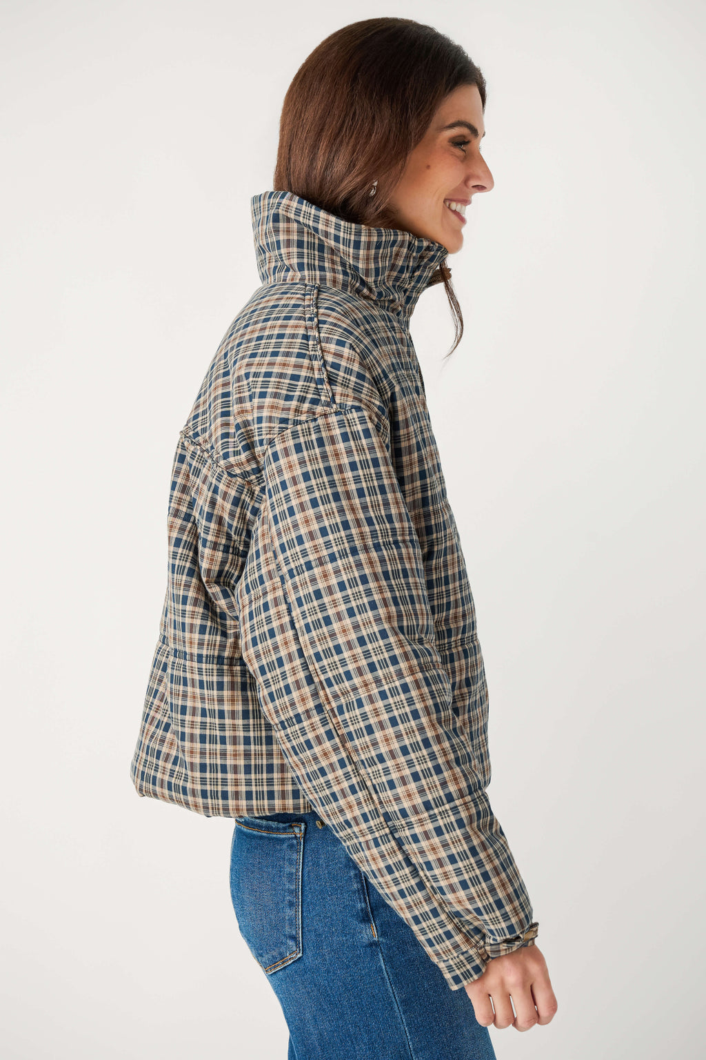 A smiling woman shown in profile wears a cropped blue and tan plaid puffer jacket with a high collar and blue jeans.