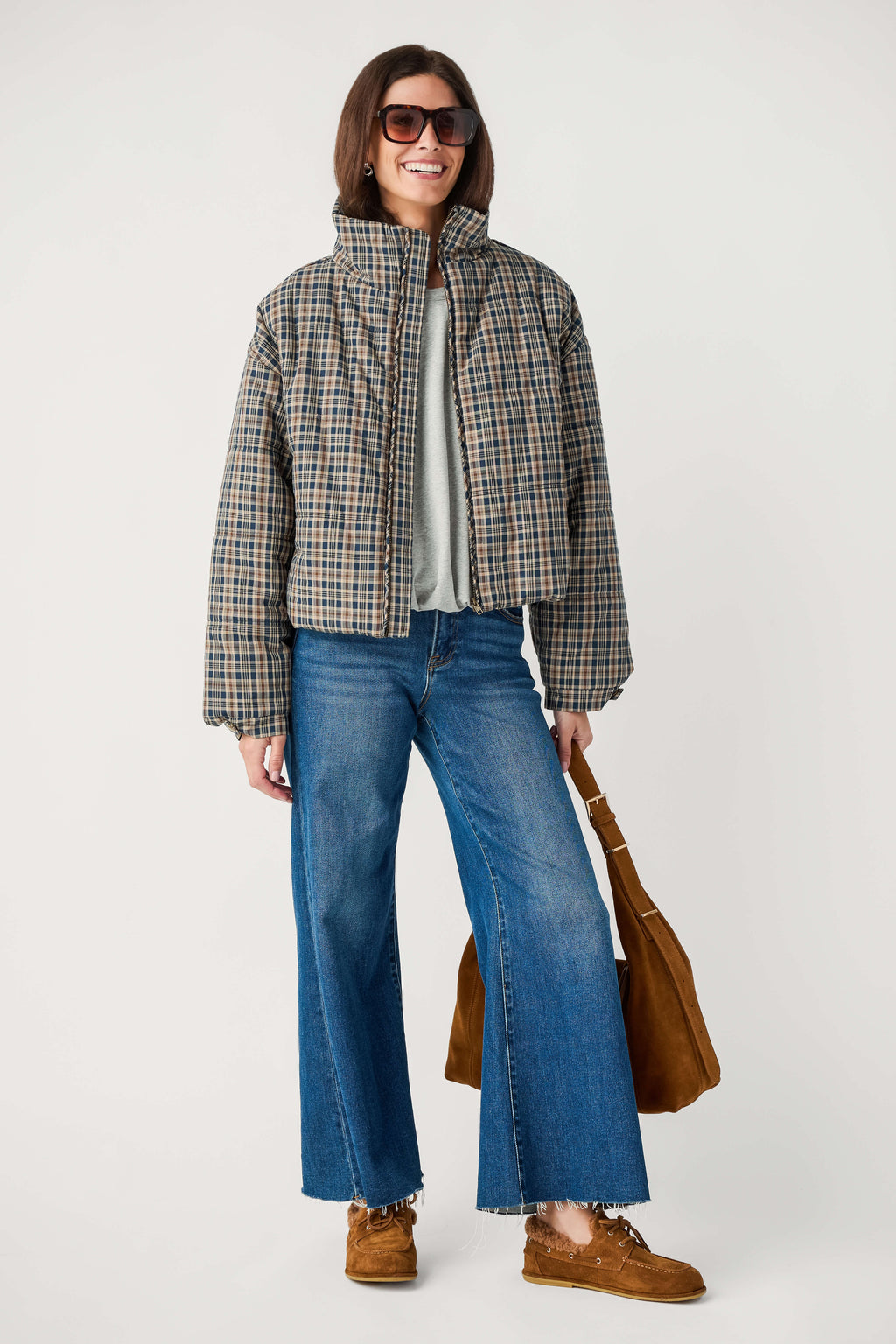 A smiling woman wears a cropped plaid puffer jacket with wide-leg jeans, tinted sunglasses, brown shoes, and a suede shoulder bag.