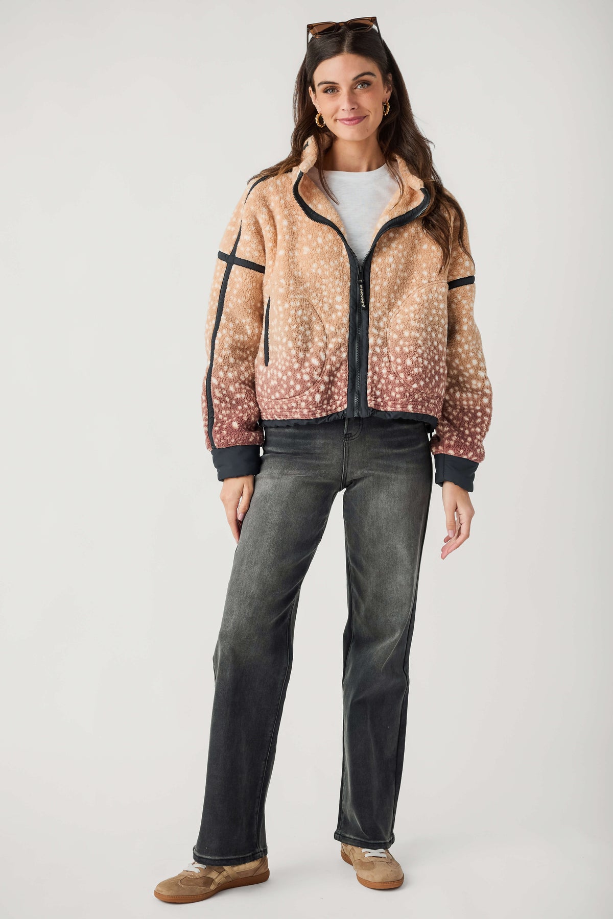 A woman modeling a zip-up fleece jacket with a beige and brown spotted pattern that fades into pinkish-brown at the hem, paired with dark gray jeans and brown shoes, against a plain light gray background.