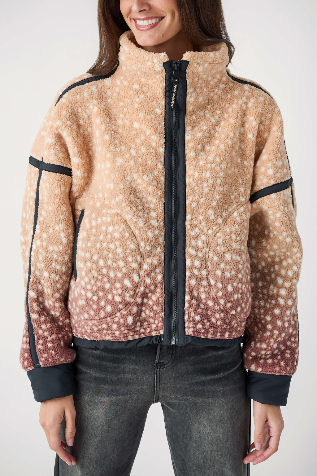 A woman wearing a sherpa fleece jacket with a beige and brown ombré deer-print pattern and black trim, paired with dark gray jeans, against a plain white studio background.