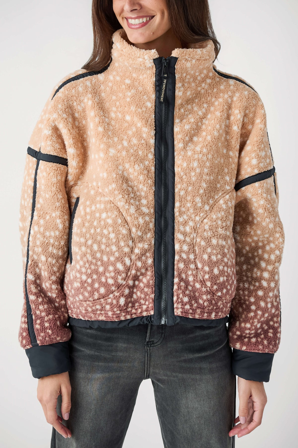 A woman wearing a sherpa fleece jacket with a beige and brown ombré deer-print pattern and black trim, paired with dark gray jeans, against a plain white studio background.