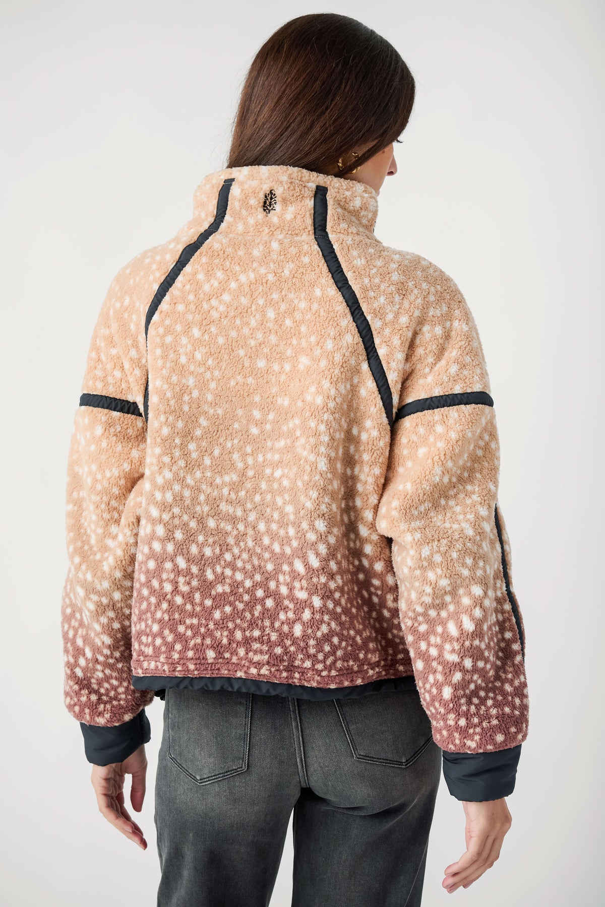 Back view of a woman wearing a tan fleece jacket with white spots, a fading berry hem, and black trim, with gray jeans.