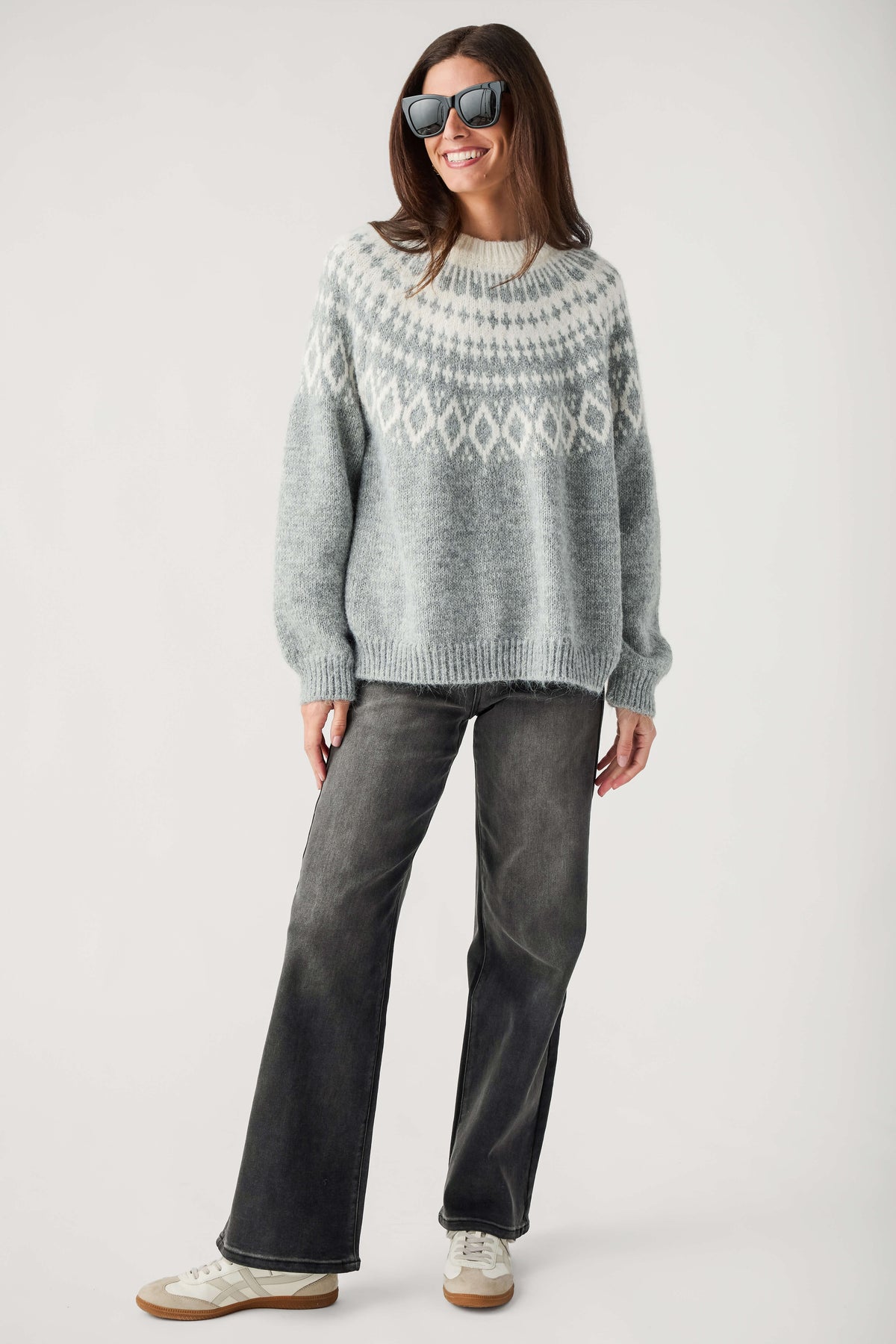A smiling woman wears a gray Fair Isle sweater with cream patterned yoke, black jeans, sunglasses, and beige sneakers.