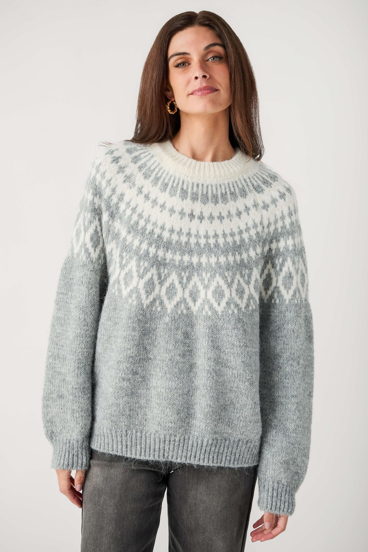 A woman wearing a light gray and white Fair Isle patterned knit sweater, paired with dark gray jeans, standing against a plain light gray studio background.