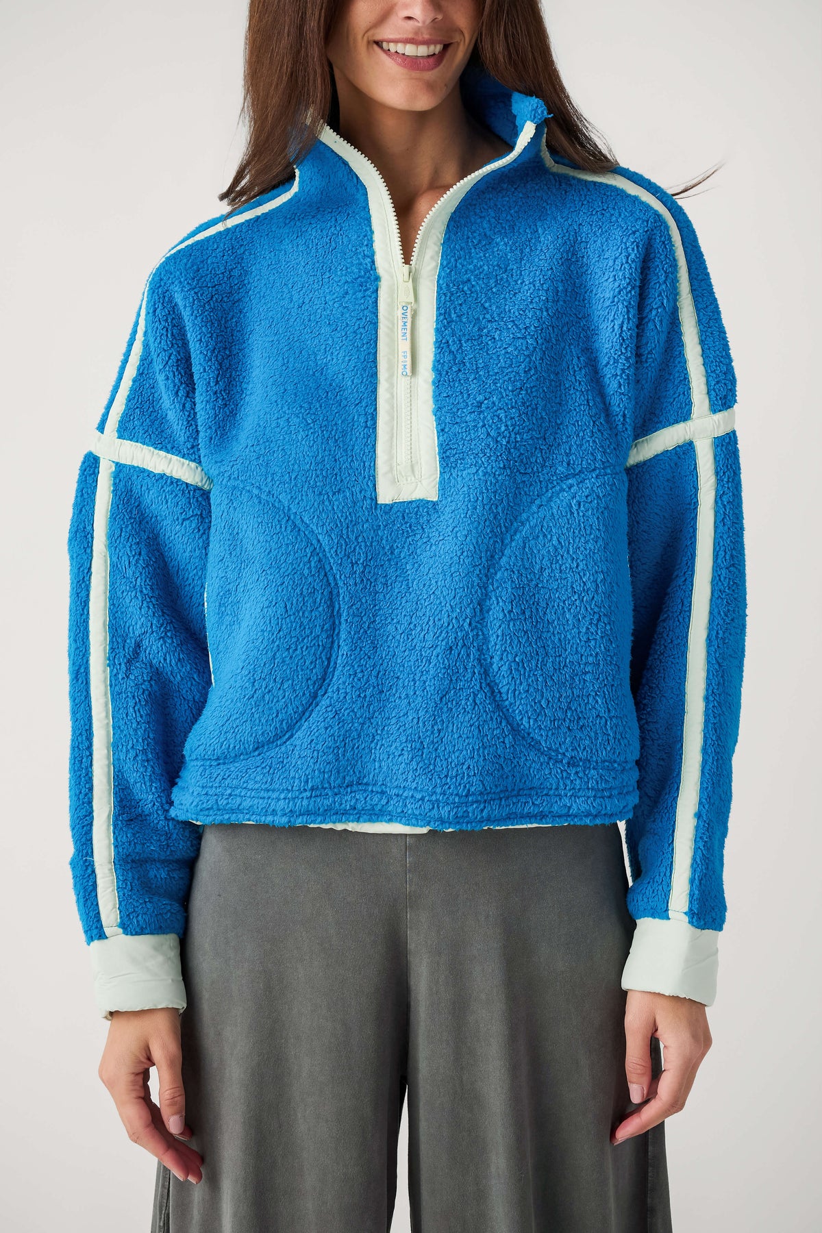 A woman wears a bright blue fleece half-zip pullover with pale mint trim and curved front pockets, paired with gray pants.