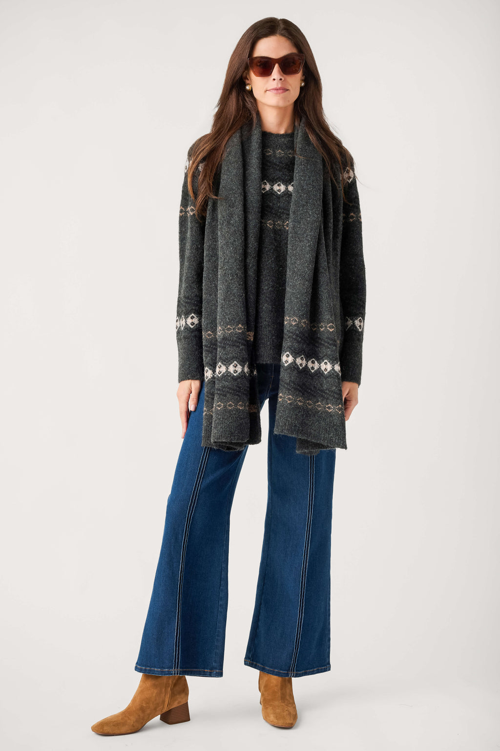 A woman wears a charcoal Fair Isle sweater with a matching long scarf, flared blue jeans, sunglasses, and tan suede ankle boots.