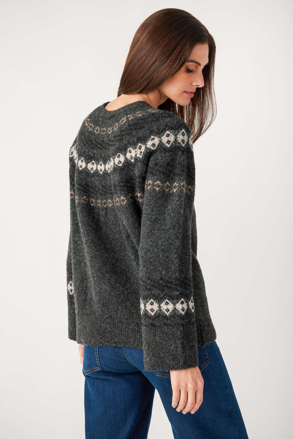 A woman with long brown hair, seen from behind, wears a charcoal Fair Isle sweater with cream patterns and blue jeans.