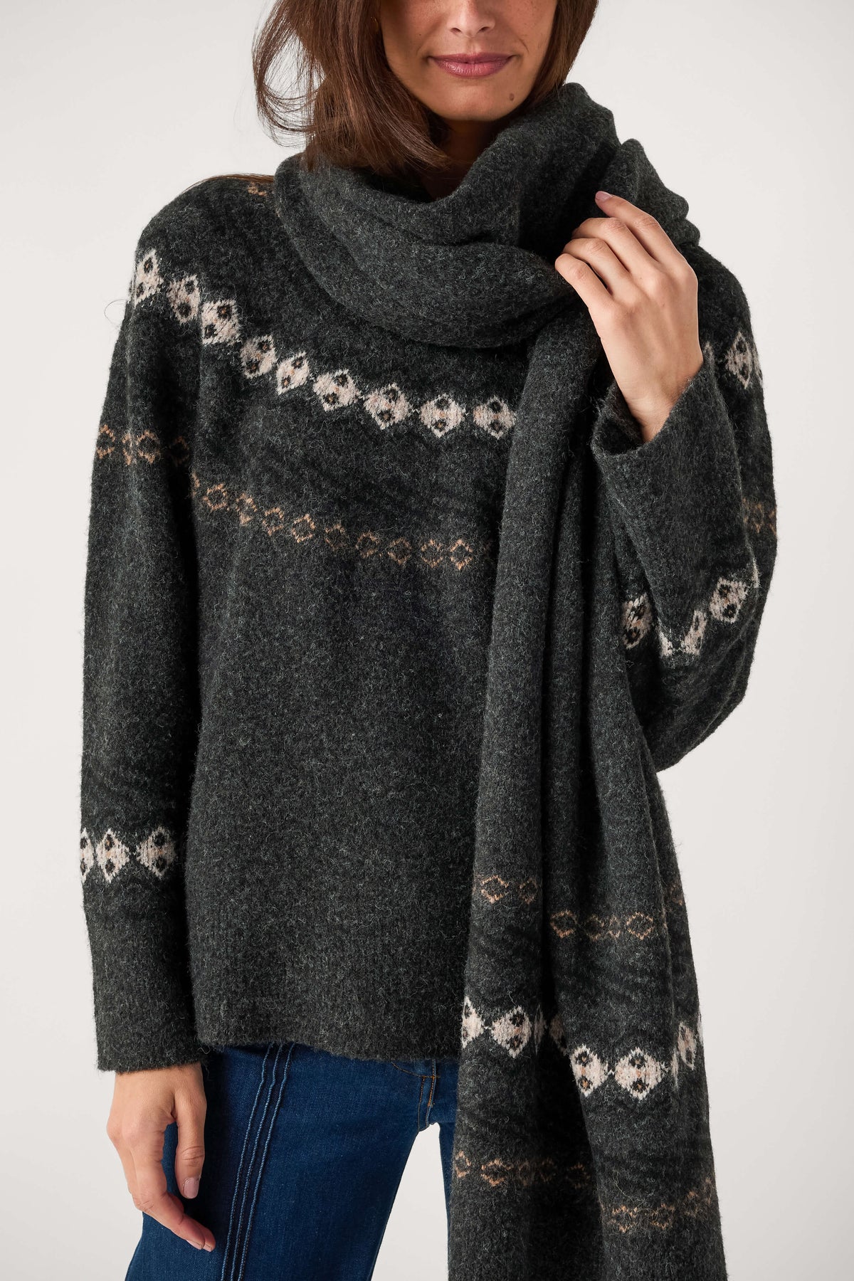 A woman wearing a dark charcoal gray knit sweater and matching scarf with cream and tan geometric Fair Isle patterns, paired with blue jeans against a plain light gray background.