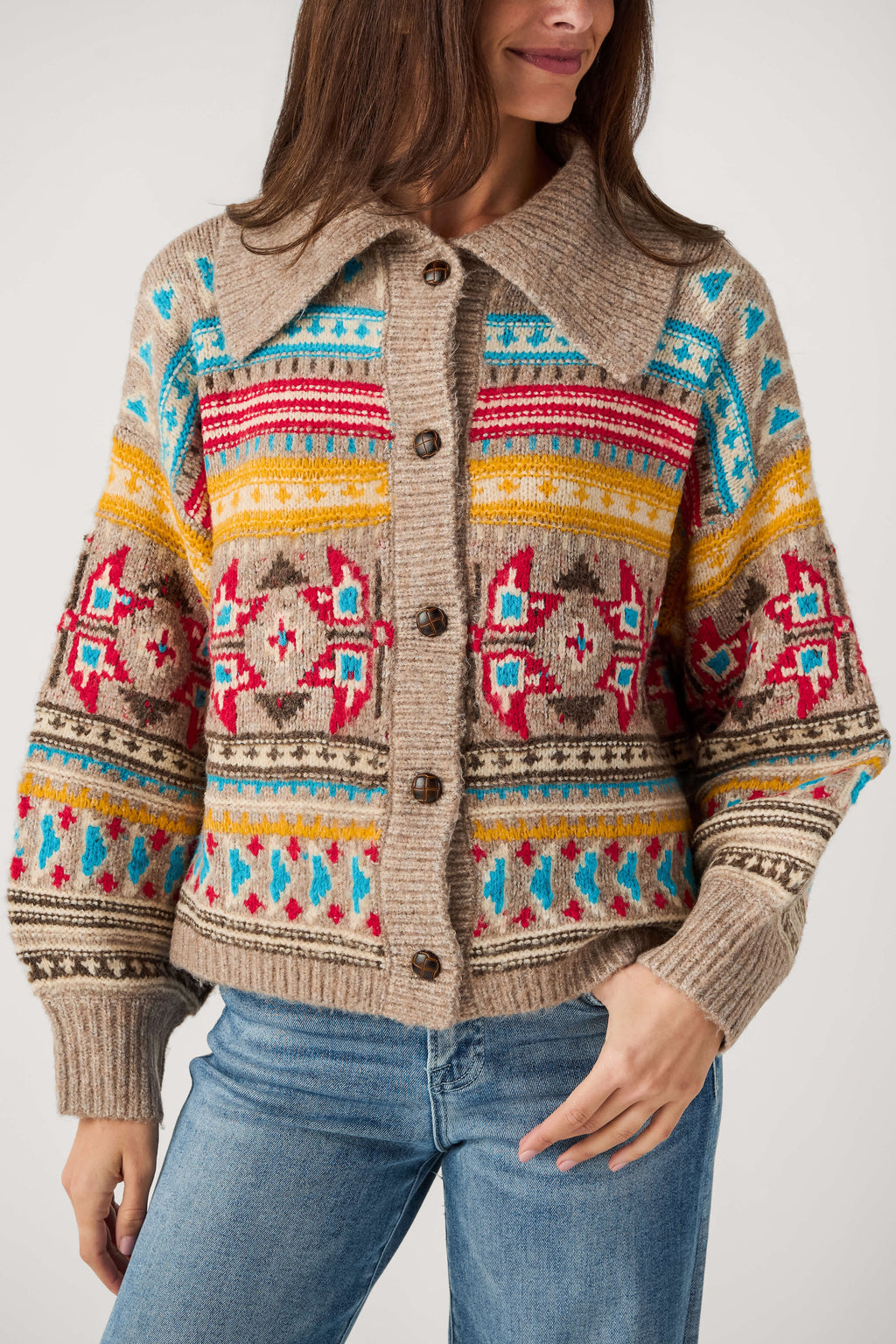A woman wears a beige knit cardigan with a wide collar, brown buttons, and red, yellow, and turquoise Fair Isle patterns over blue jeans.