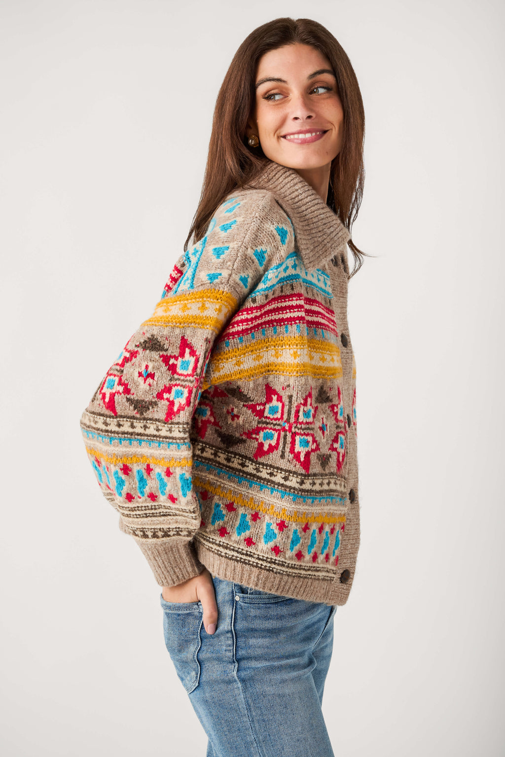 A smiling woman wears a beige button-up cardigan with red, blue, and yellow Fair Isle patterns and light blue jeans.