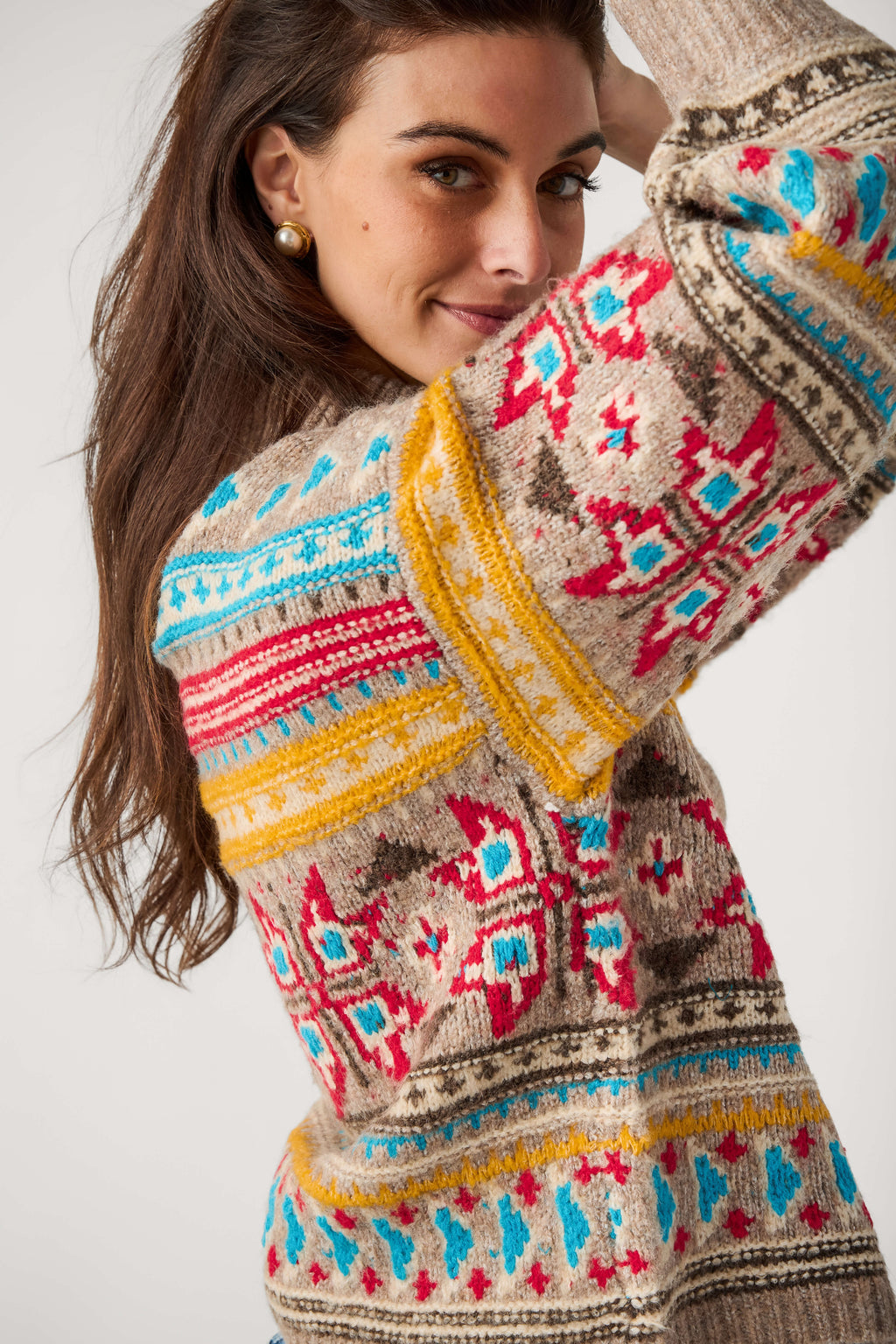 A woman wearing a beige knit sweater with a colorful Fair Isle pattern in red, blue, yellow, and brown, photographed against a light gray studio background.