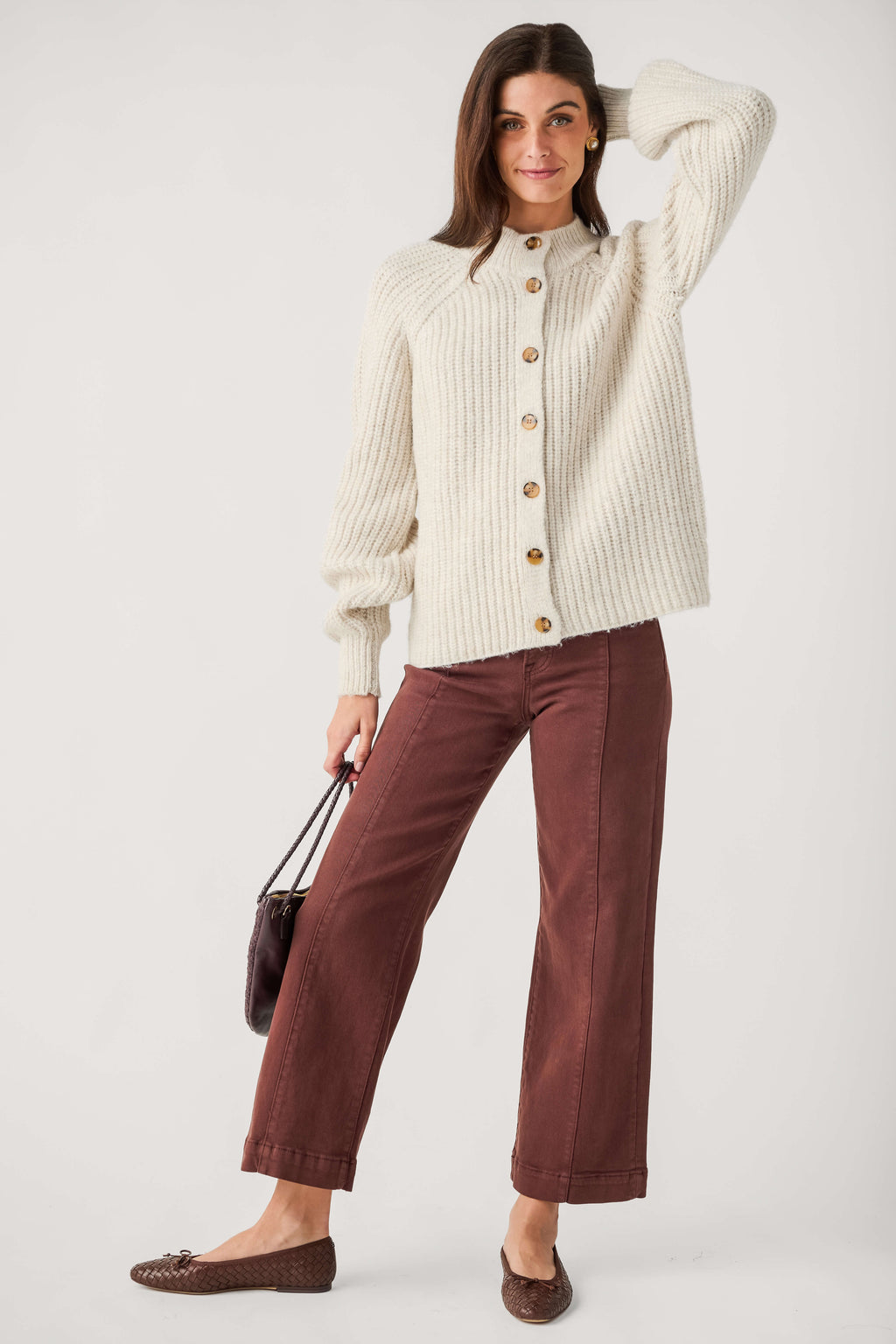 A woman wears a cream ribbed mock neck cardigan with brown buttons, brown wide-leg pants, and woven flats while holding a handbag.