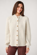 A smiling woman wears a cream ribbed knit cardigan with a mock neck, tortoiseshell buttons, and full sleeves with banded cuffs.