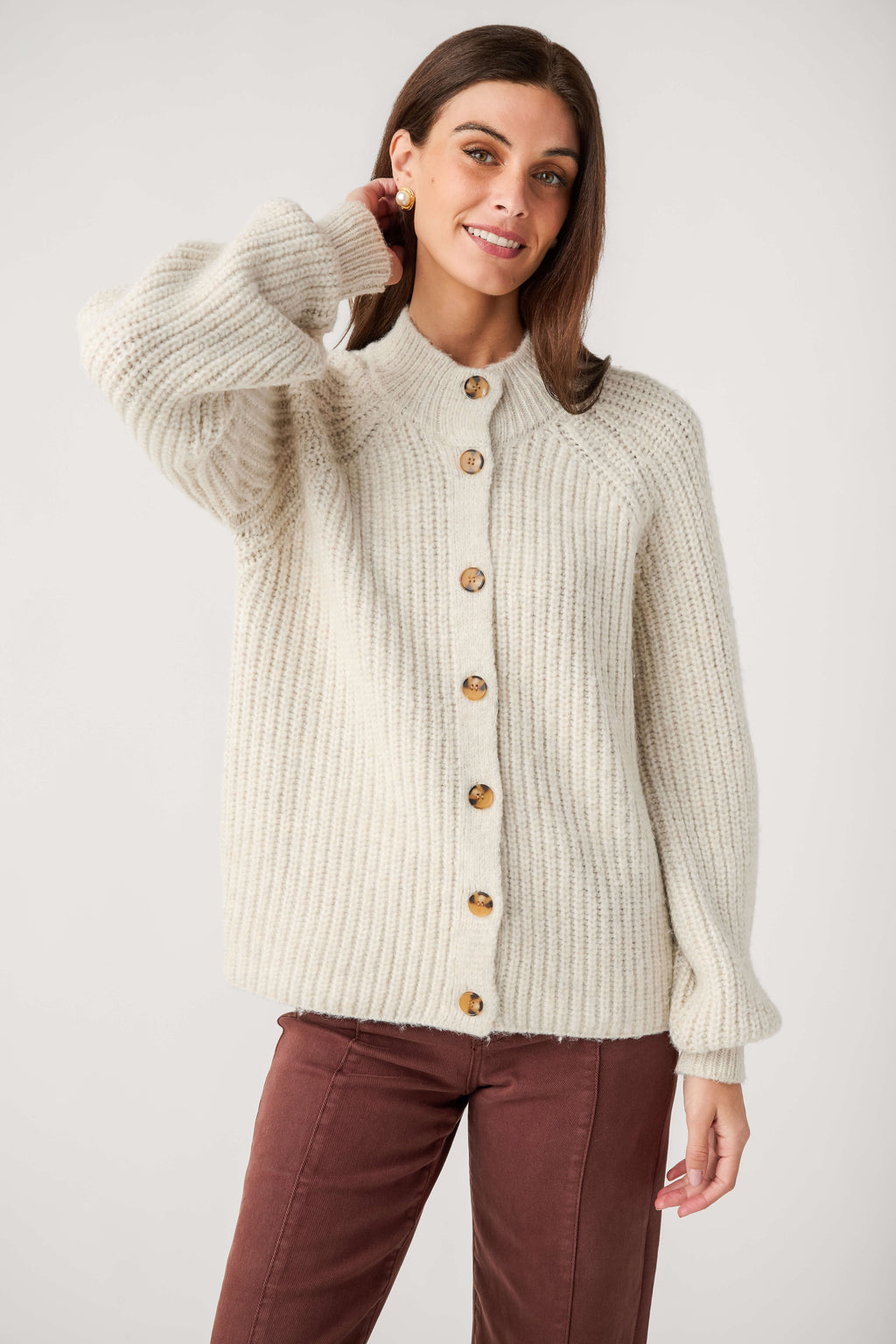 A woman wears a cream ribbed knit cardigan with tortoiseshell buttons, a mock neck, and balloon sleeves, paired with brown pants.