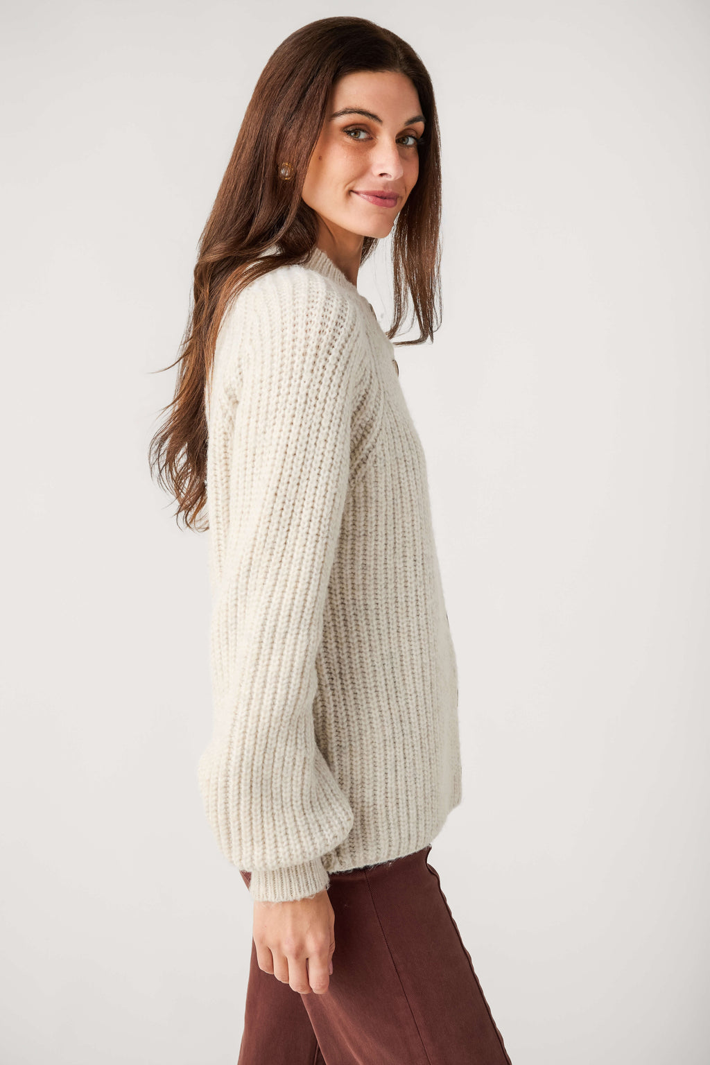 A woman with long brown hair wears a cream ribbed knit mock neck cardigan with buttons and brown pants, shown from the side.