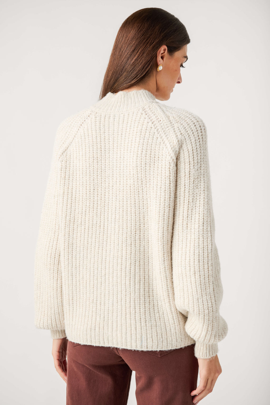 Back view of a woman wearing a cream chunky ribbed mock neck cardigan with full sleeves and brown pants.