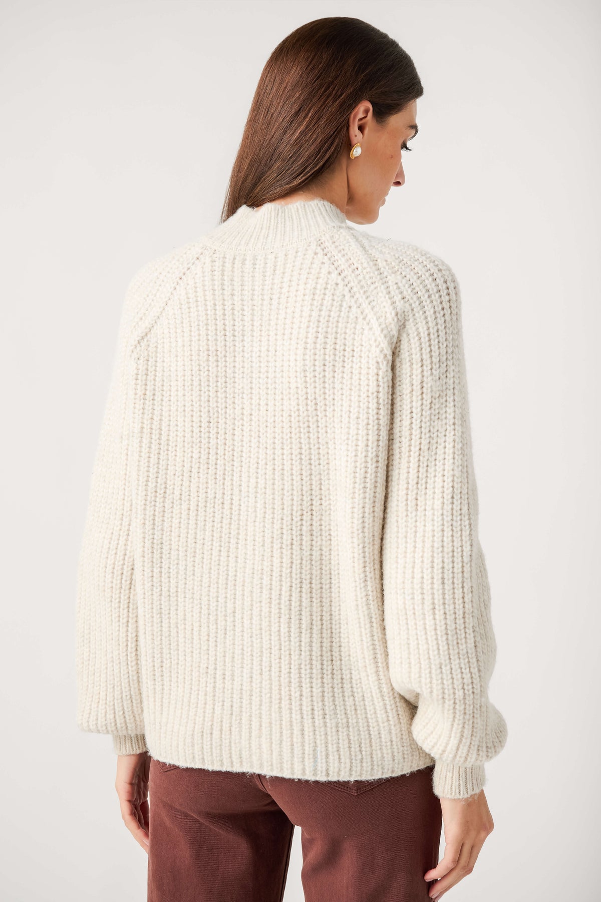 Back view of a woman wearing a cream chunky ribbed mock neck cardigan with full sleeves and brown pants.