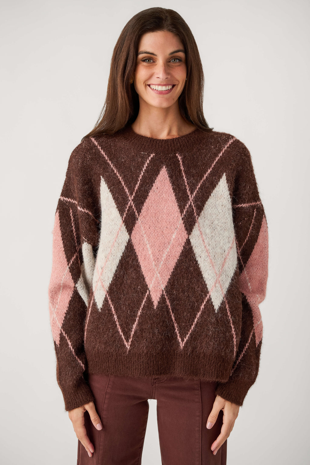 A smiling woman with long brown hair wears a fuzzy brown crewneck sweater with pink and cream argyle diamonds and matching brown pants.
