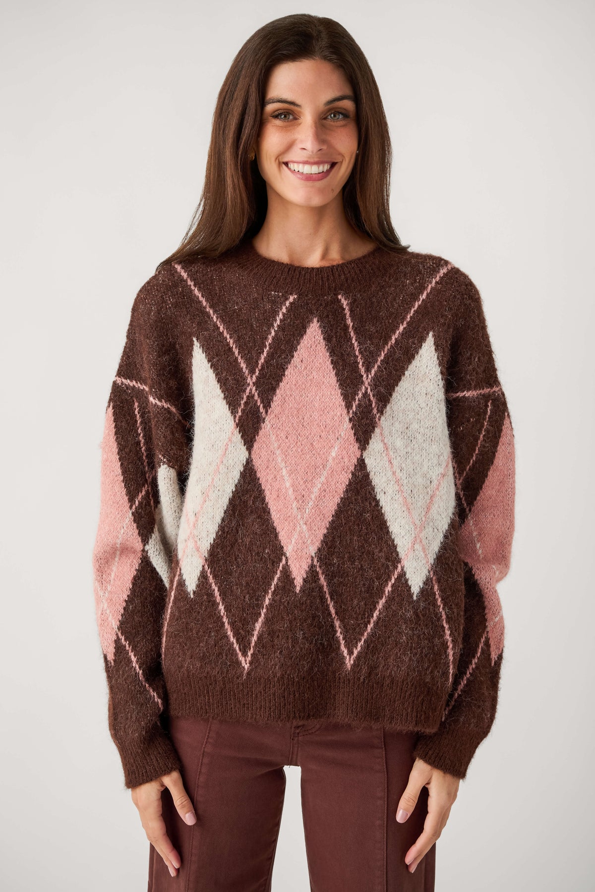 A smiling woman with long brown hair wears a fuzzy brown crewneck sweater with pink and cream argyle diamonds and matching brown pants.