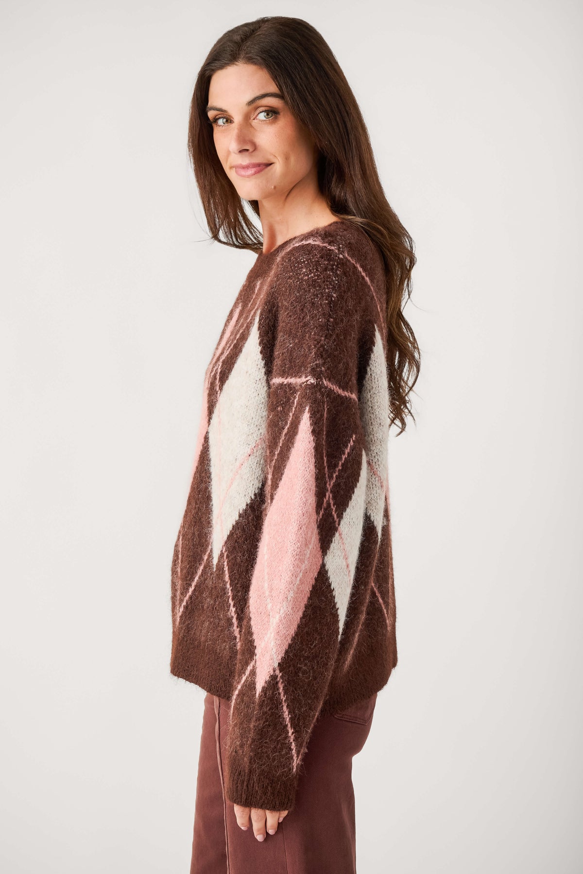 A woman with long brown hair wears a fuzzy brown crewneck sweater with pink and cream argyle diamonds and maroon pants.