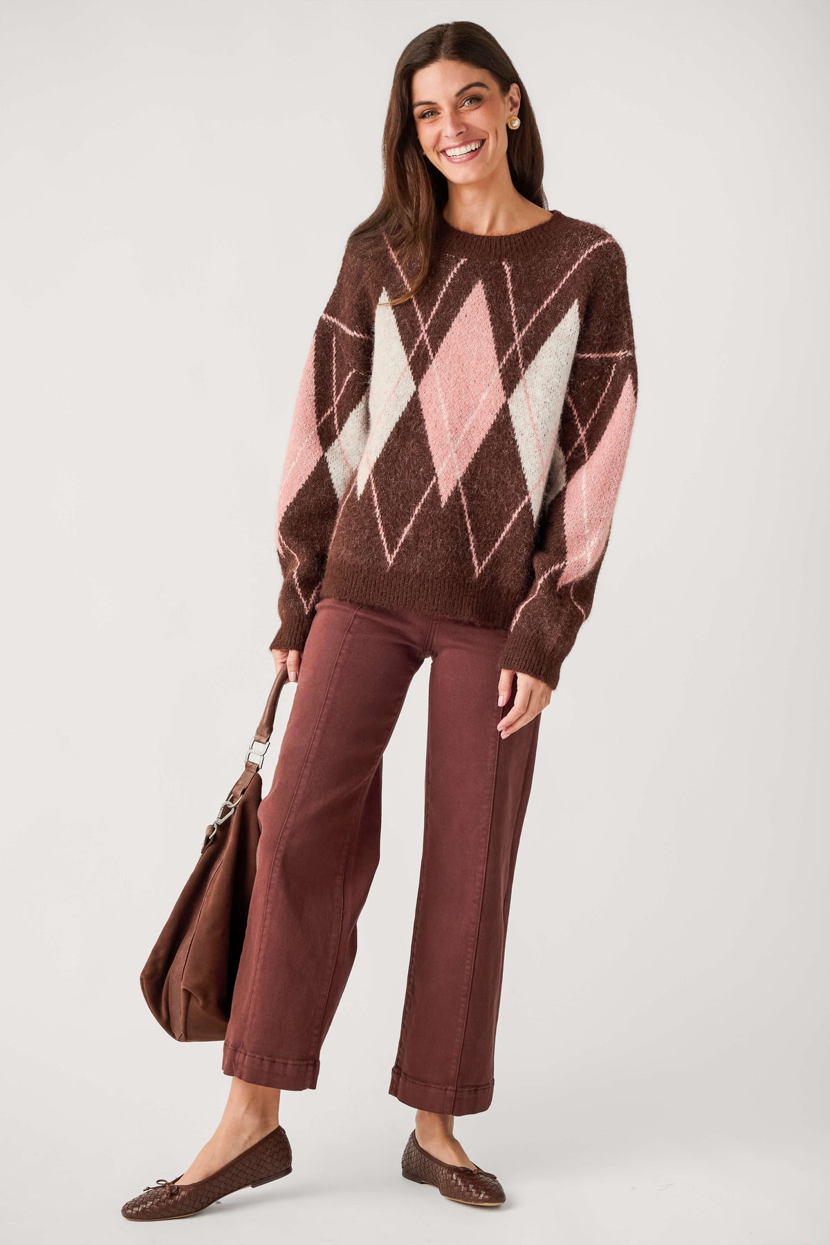 A woman modeling a brown knit sweater with a pink and cream argyle pattern, paired with rust-colored wide-leg pants, brown flats, and a brown tote bag, against a light gray studio background.