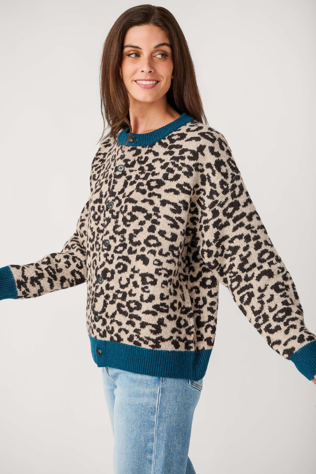 A smiling woman wears a button-front cardigan with beige and black leopard print, teal ribbed trim, and light blue jeans.